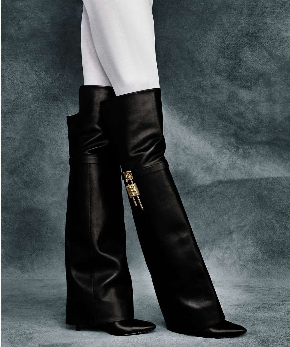 A person wearing white pants and black leather over-the-knee boots with gold-colored hardware, posing in a fashion studio.