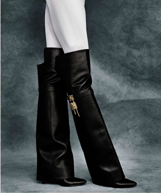 A person wearing white pants and black leather over-the-knee boots with gold-colored hardware, posing in a fashion studio.