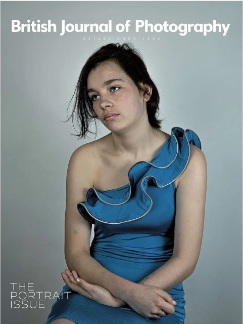 A young woman with dark hair and fair skin wearing a blue, one-shoulder dress, sits against a grey background on the British Journal of Photography cover.