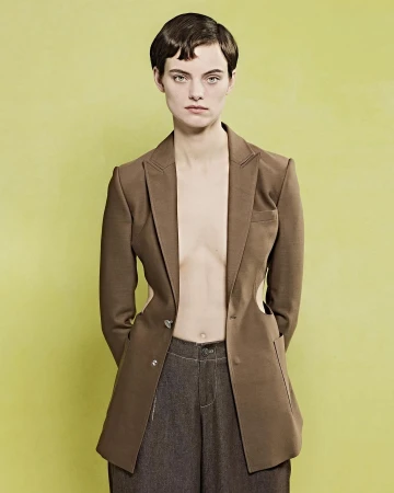 Model with short dark hair wearing an open brown blazer and dark trousers against a lime green background.