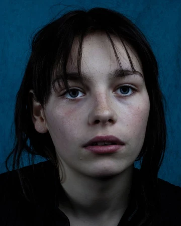 Close-up portrait of a young woman with dark hair and blue eyes against a textured dark blue background.