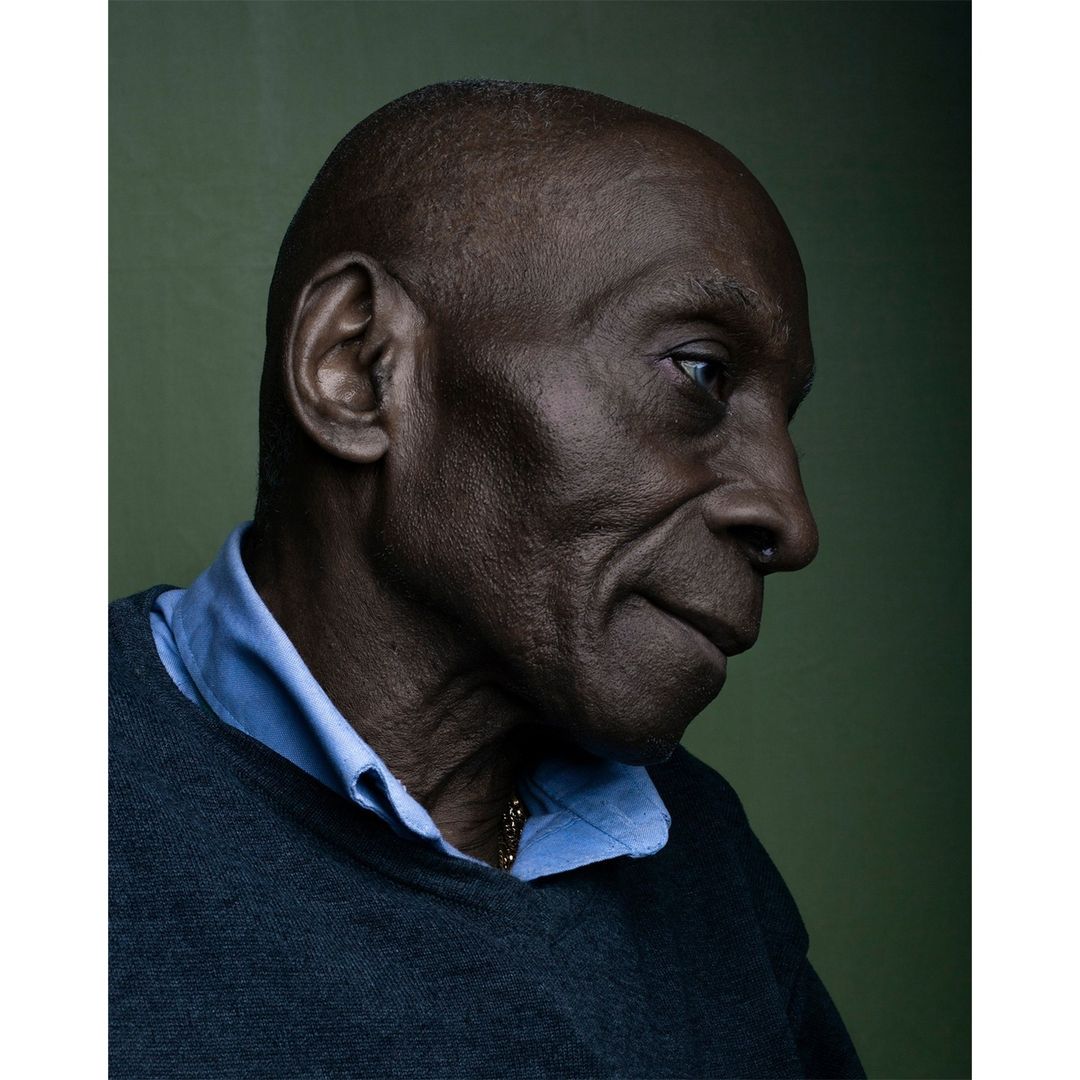 Close-up portrait of an adult Black man with a shaved head, wearing a blue collared shirt and gray sweater, set against an olive green background.