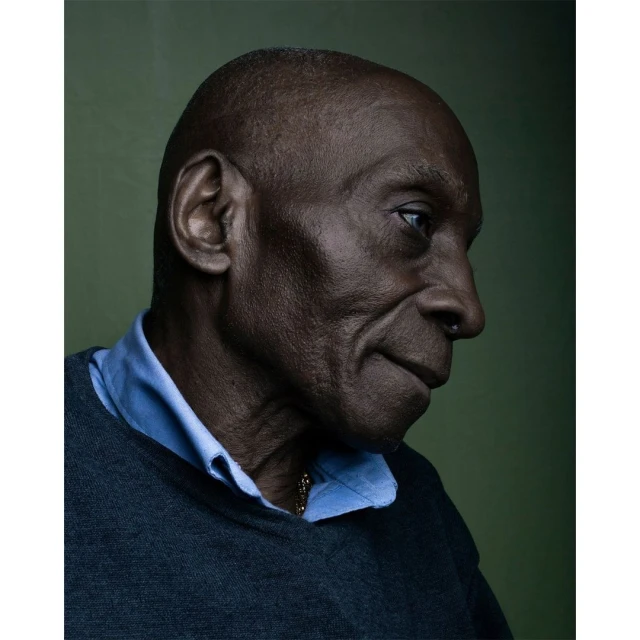Close-up portrait of an adult Black man with a shaved head, wearing a blue collared shirt and gray sweater, set against an olive green background.