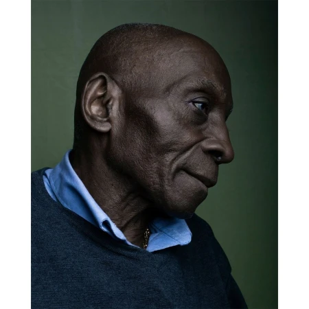 Close-up portrait of an adult Black man with a shaved head, wearing a blue collared shirt and gray sweater, set against an olive green background.