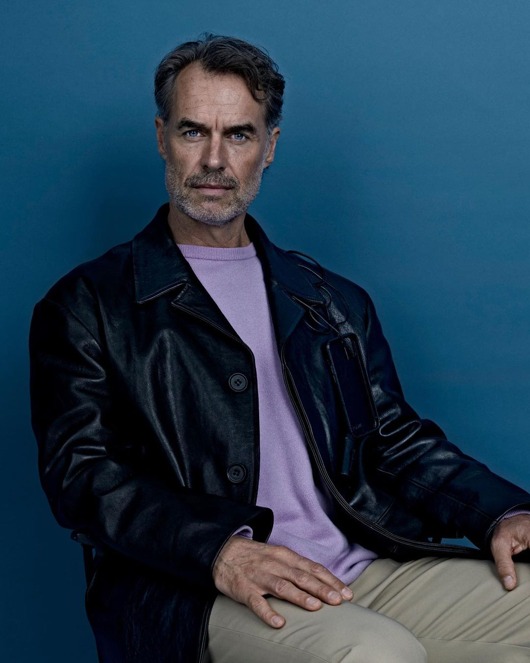 Portrait of a mature man in a leather jacket, light purple sweater, and khaki pants against a blue background looking at the camera.