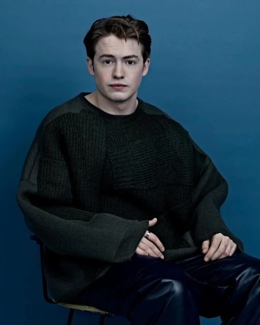 Portrait of a young adult male with light brown hair wearing a dark green sweater and dark pants, seated against a blue background.