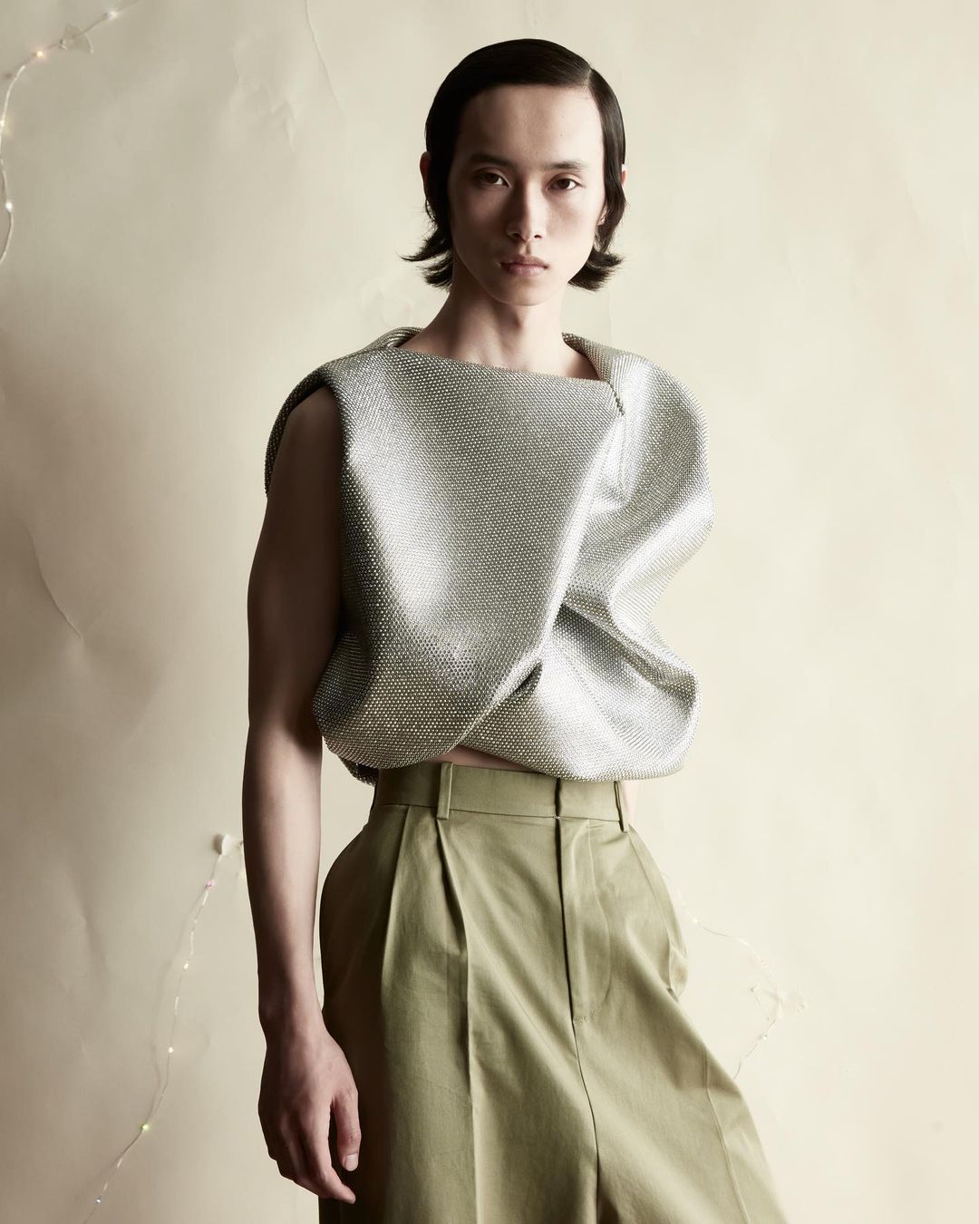 Asian male model with slicked-back hair poses in a silver textured top and pleated olive-green pants against a neutral backdrop.