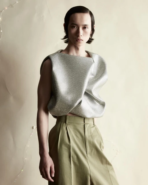 Asian male model with slicked-back hair poses in a silver textured top and pleated olive-green pants against a neutral backdrop.