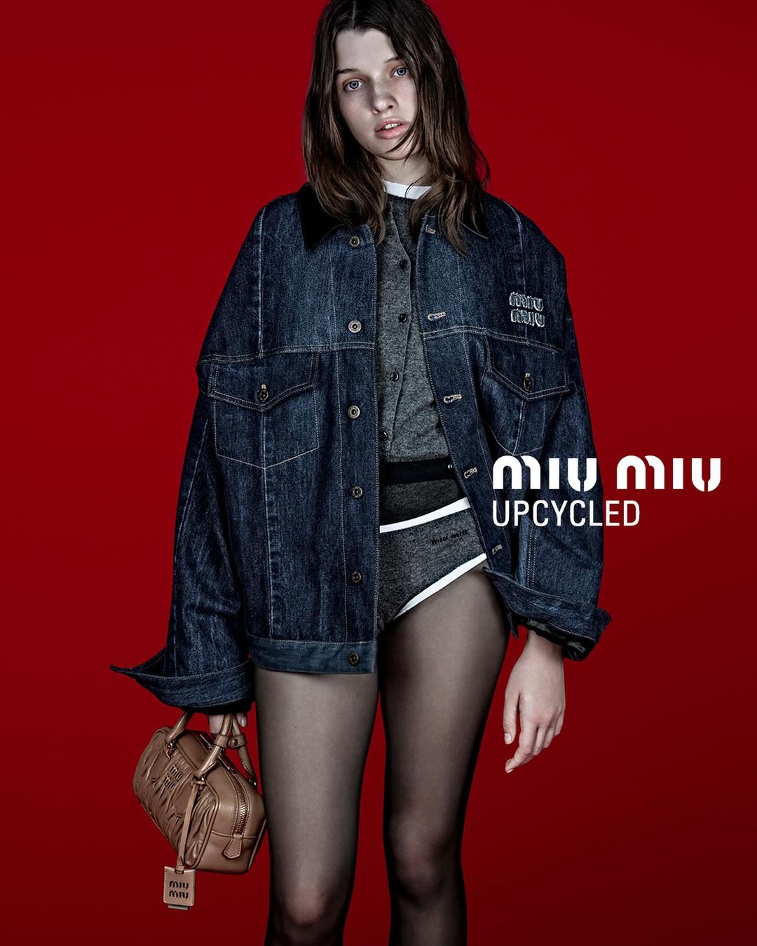 Model wearing an oversized denim jacket and carrying a brown leather handbag against a solid red background
