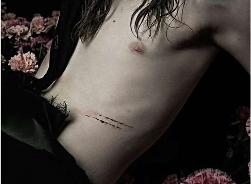 Close-up studio shot of a shirtless person with long brown hair, and floral accents, against a black background. Fashion editorial style.
