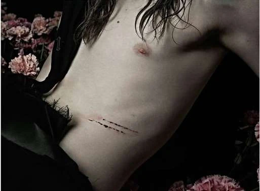 Close-up studio shot of a shirtless person with long brown hair, and floral accents, against a black background. Fashion editorial style.