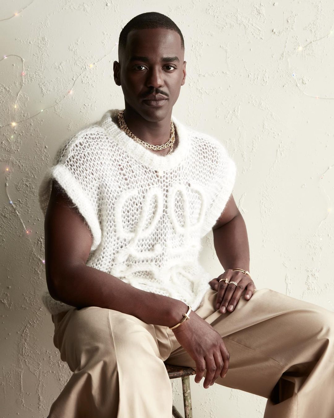 Portrait of adult male with a mustache wearing a white knit top and gold jewelry, seated against an off-white textured wall.