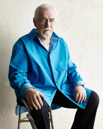 Portrait of Brian Cox seated on a stool wearing a bright blue shirt and dark trousers against a textured off-white wall