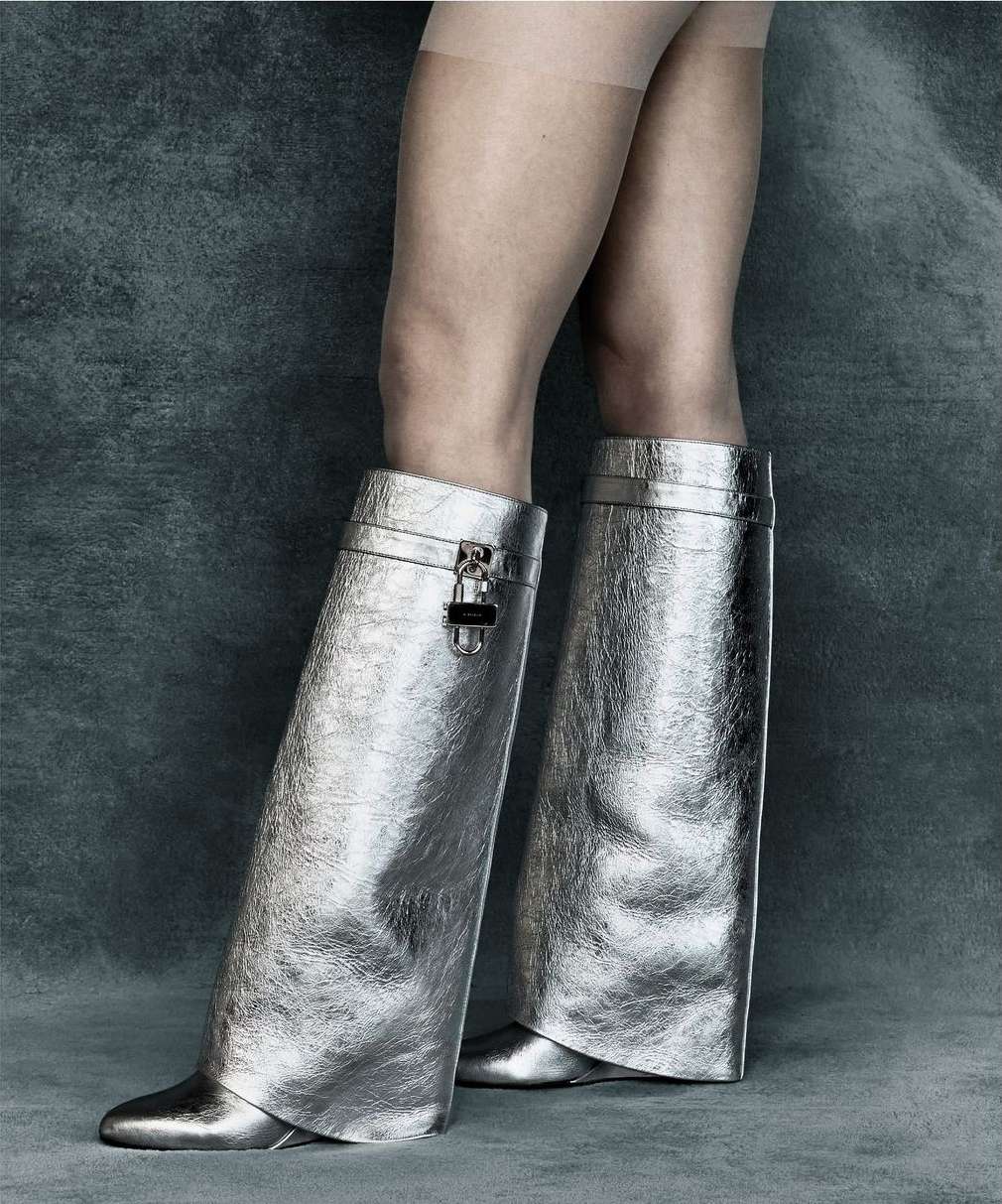 A person wearing metallic silver, high-fashion, above-the-ankle, slouchy boots with a pointed toe, against a mottled gray background.