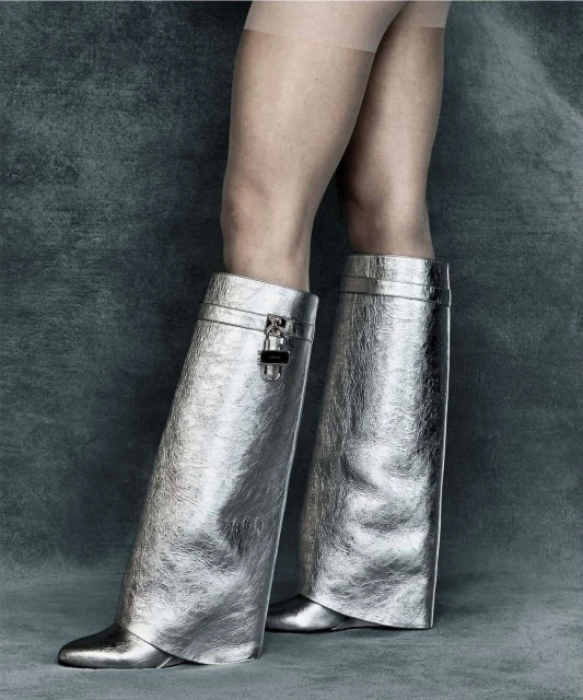 A person wearing metallic silver, high-fashion, above-the-ankle, slouchy boots with a pointed toe, against a mottled gray background.