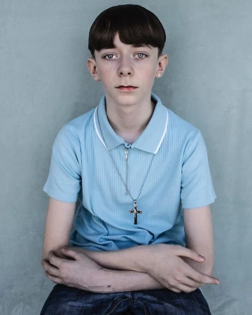 Close-up portrait of a fair-skinned young boy with a bowl cut hairstyle, wearing a blue polo shirt and a silver cross necklace indoors.