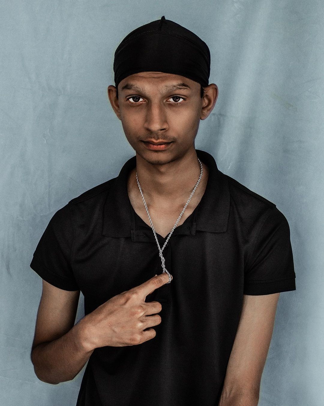 Portrait of a young adult male wearing a black durag and shirt, silver chain with a pendant, pointing to it with a neutral expression against a grey backdrop.