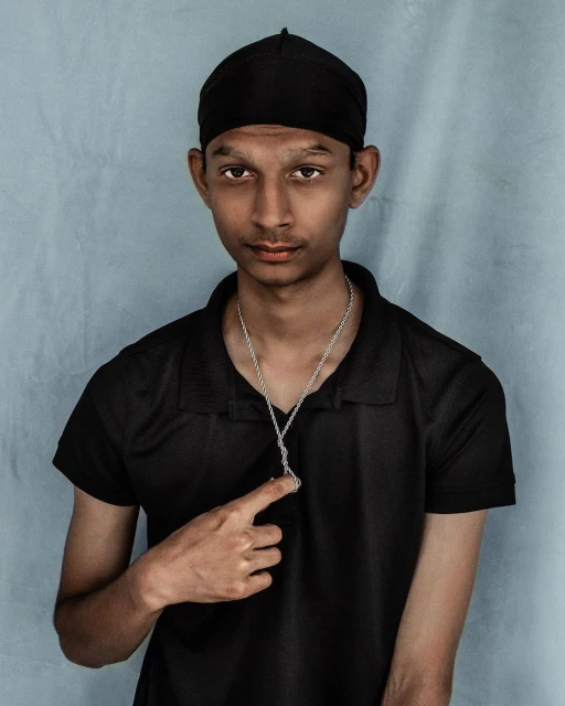 Portrait of a young adult male wearing a black durag and shirt, silver chain with a pendant, pointing to it with a neutral expression against a grey backdrop.