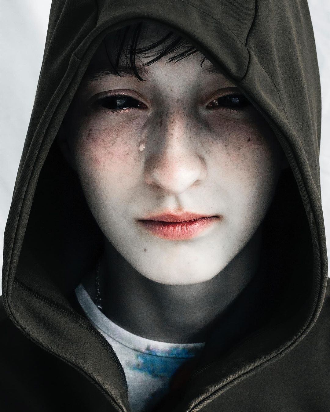 Portrait of a young person with dark eyes and a hoodie, a tear visible on their face, against a bright background.