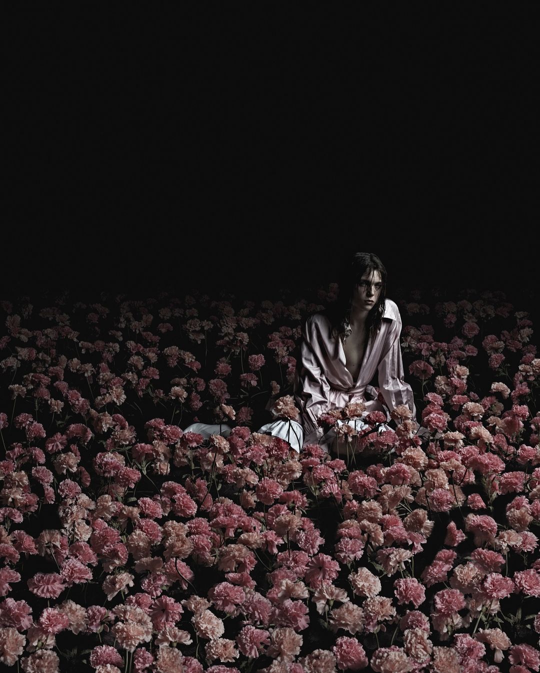 Person in unbuttoned shirt seated in a field of pink flowers in the dark with a high-contrast exposure and low eye-level angle.