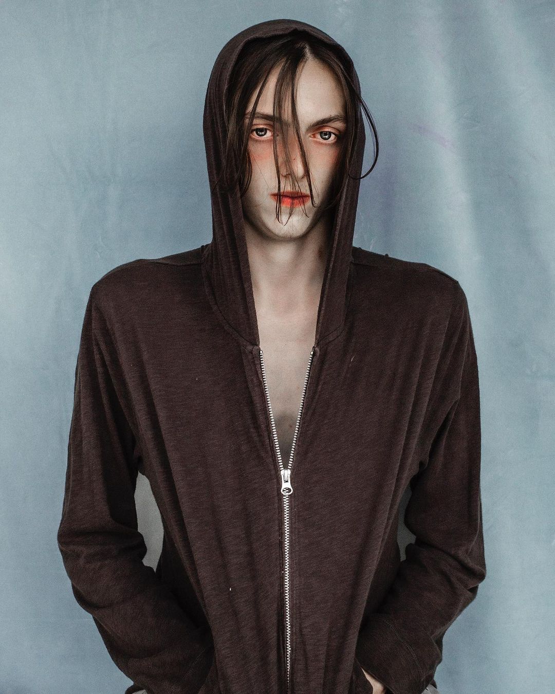 Portrait of a person wearing a brown hooded zip-up sweatshirt against a textured pale blue background by Jet Swan.