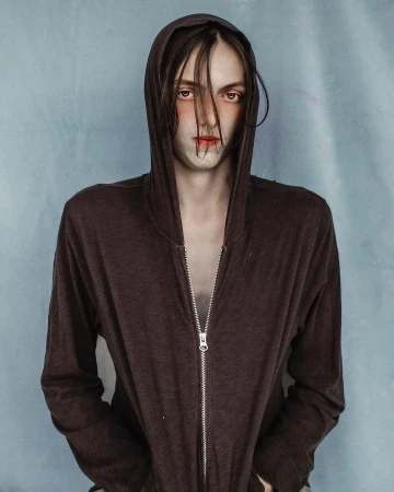Portrait of a person wearing a brown hooded zip-up sweatshirt against a textured pale blue background by Jet Swan.
