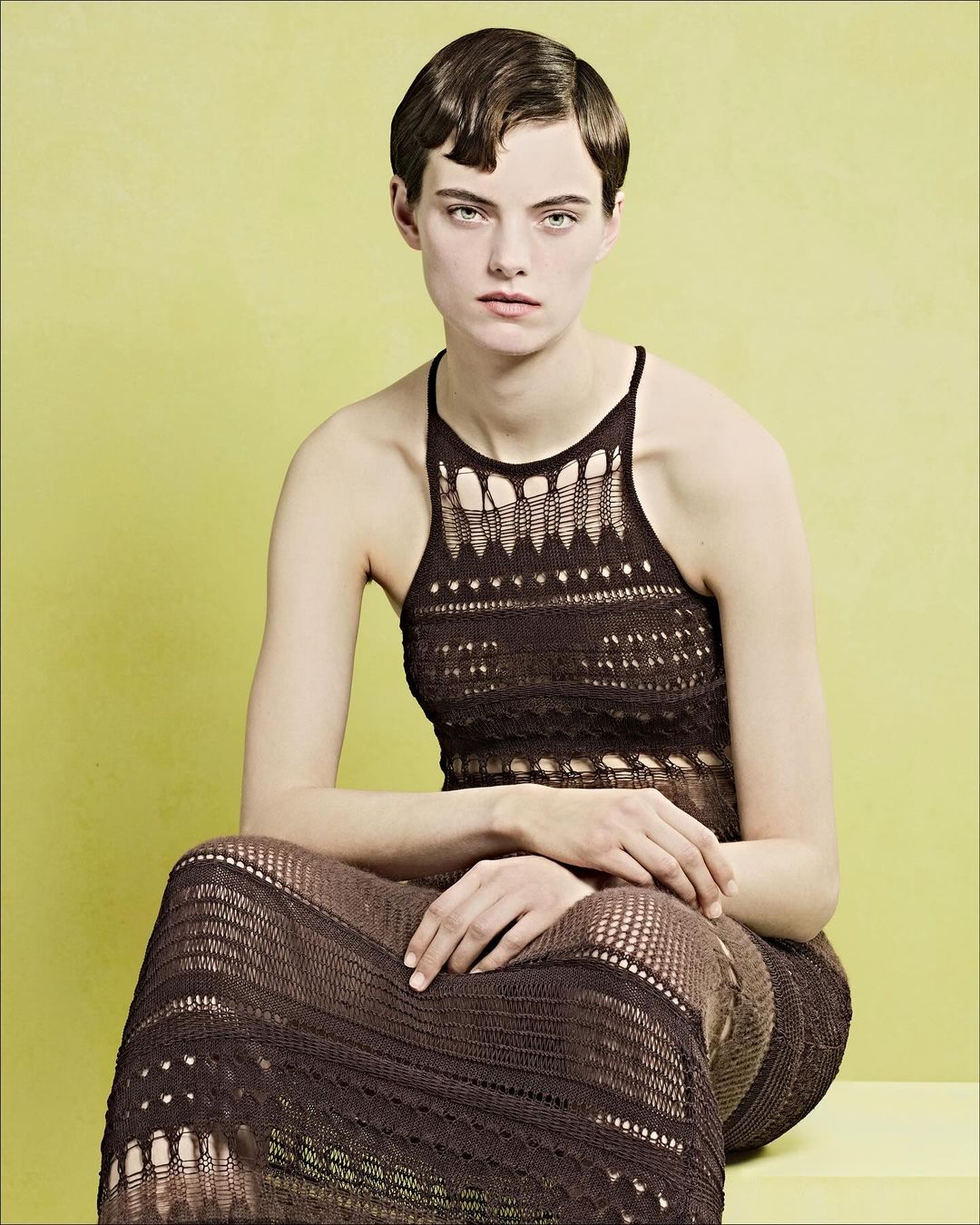 Model with short styled hair wearing a brown knit dress seated against a solid yellow-green background