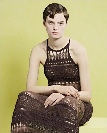 Model with short styled hair wearing a brown knit dress seated against a solid yellow-green background
