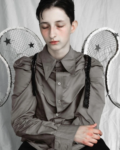 Young person with pale skin, wearing a button-down shirt with ruffled sleeves, and sporting tennis racket wings, poses against a white backdrop.