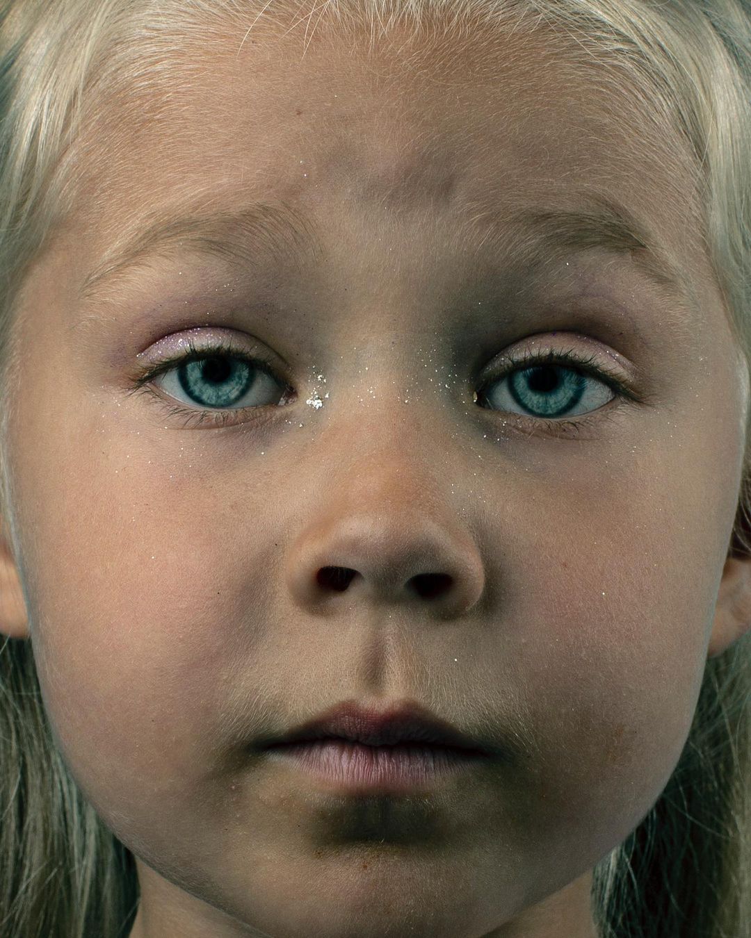 Close-up portrait of a fair-skinned child with bright blue eyes, light blonde hair, light pink eye shadow and light dusting of glitter.