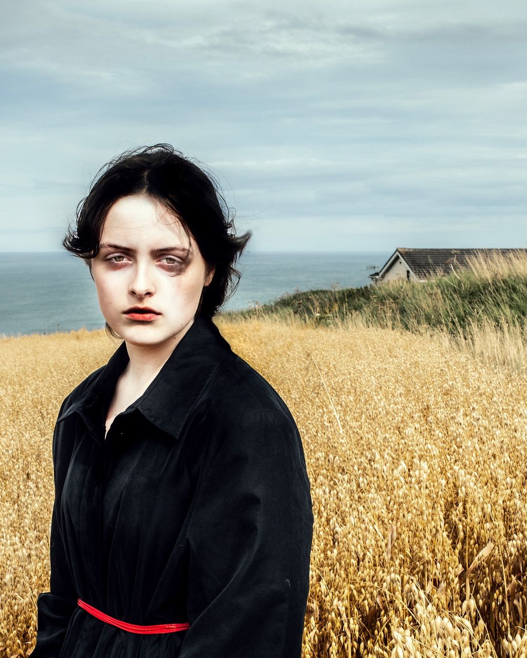 A young woman in a black coat stands in a golden field near the ocean under a cloudy sky. She has dark hair and light skin.