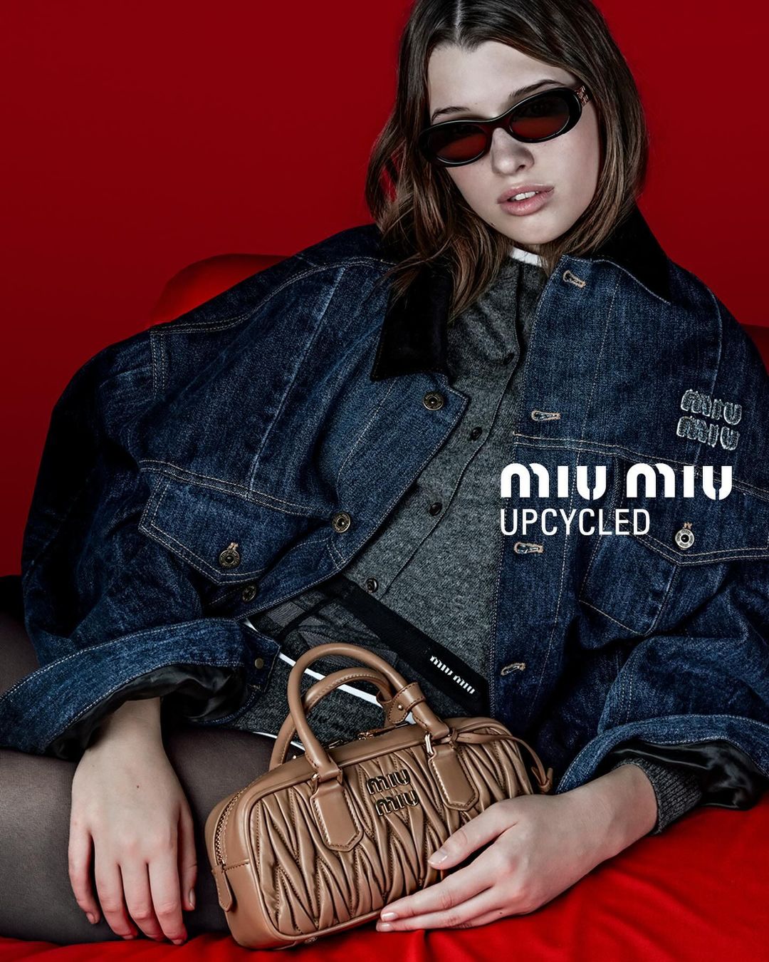Brunette woman in sunglasses and denim jacket holds a tan leather purse against a red background in a fashion studio shot.