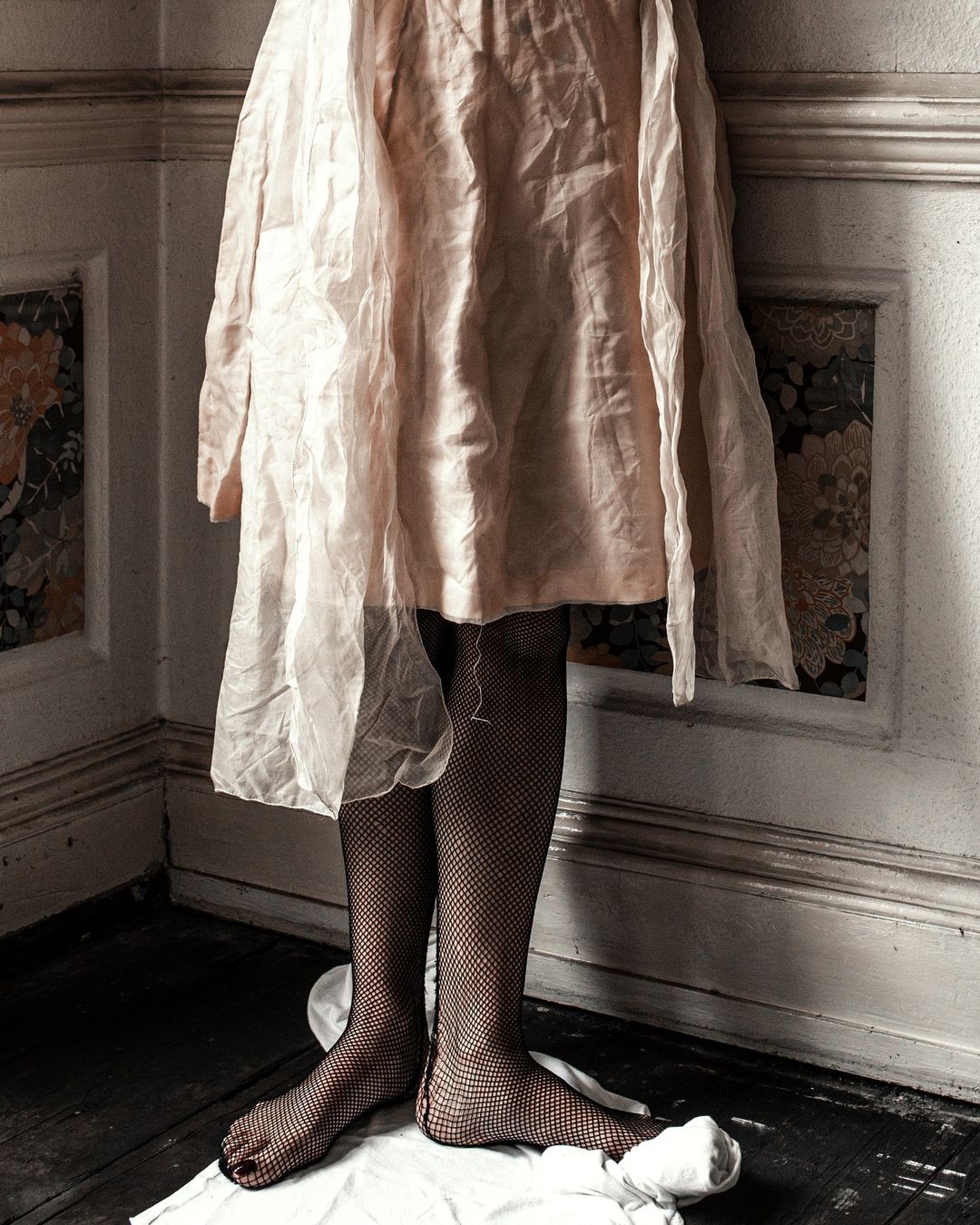 Partial view of a person in a light pink dress and fishnet stockings, standing barefoot on a white cloth in a corner of a room.