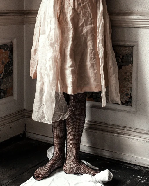 Partial view of a person in a light pink dress and fishnet stockings, standing barefoot on a white cloth in a corner of a room.