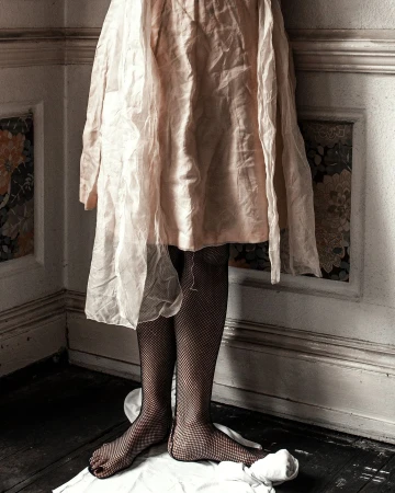 Partial view of a person in a light pink dress and fishnet stockings, standing barefoot on a white cloth in a corner of a room.