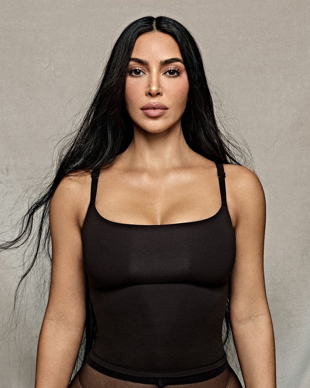 Portrait of Kim Kardashian with long dark hair wearing a black square neckline top against a textured grey wall background
