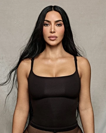 Portrait of Kim Kardashian with long dark hair wearing a black square neckline top against a textured grey wall background