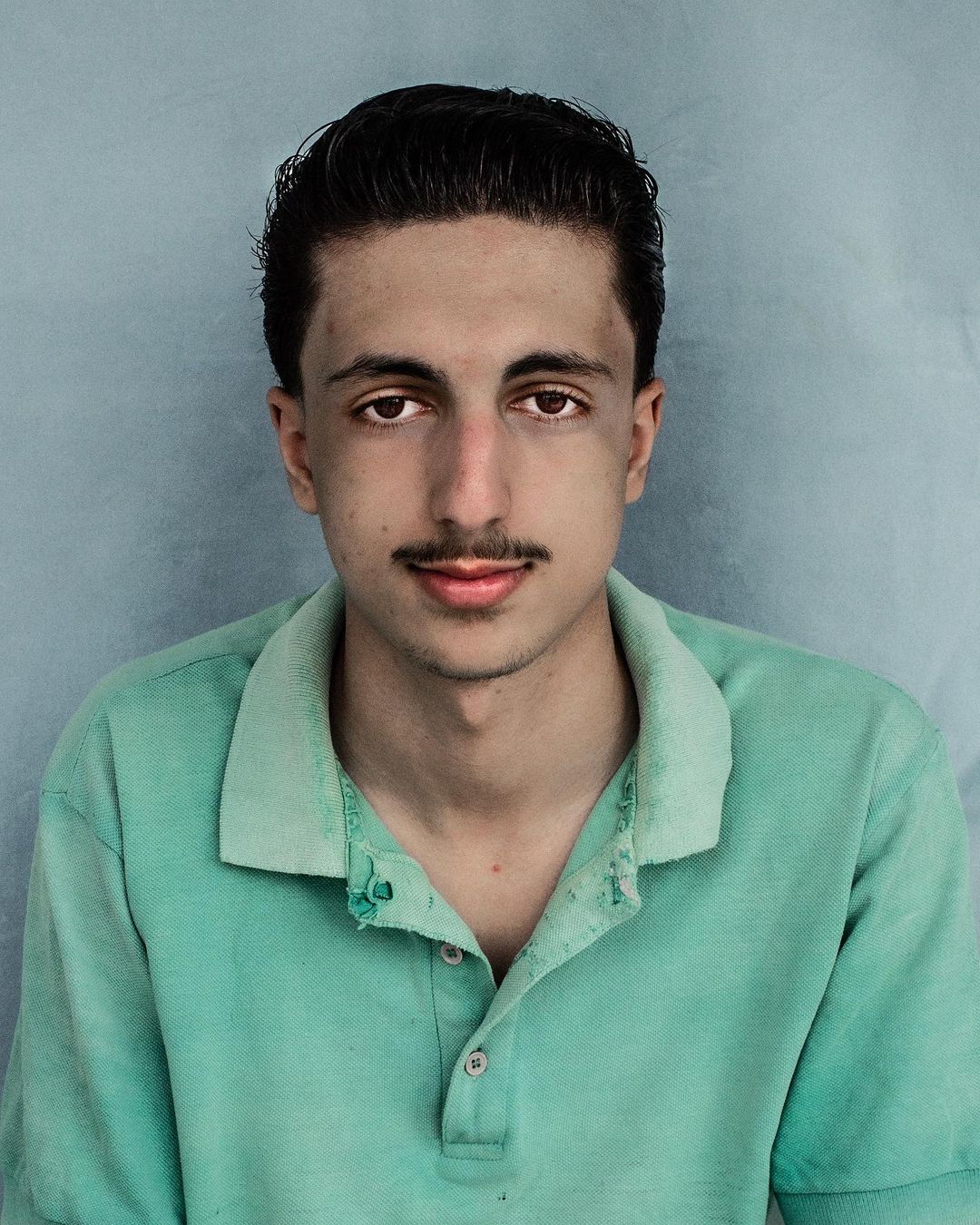 Close-up portrait of a young adult male with brown hair and a mustache, wearing a green polo shirt against a textured blue background.