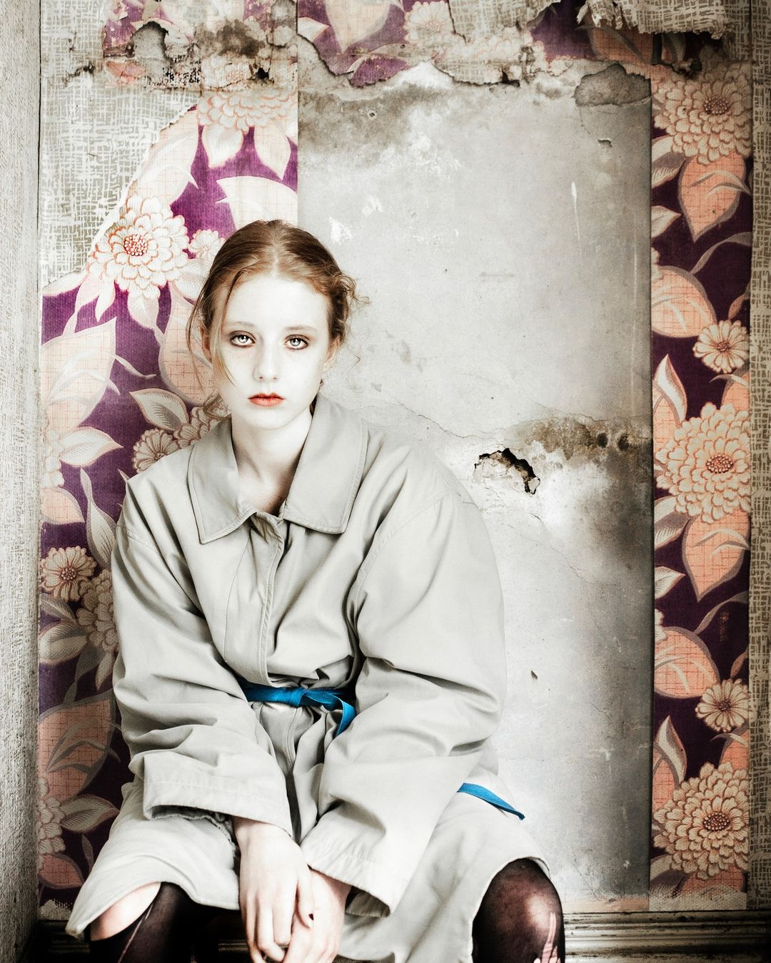 A young adult female in a grey trench coat and tights sits indoors against old wallpaper in a full shot, facing the camera.