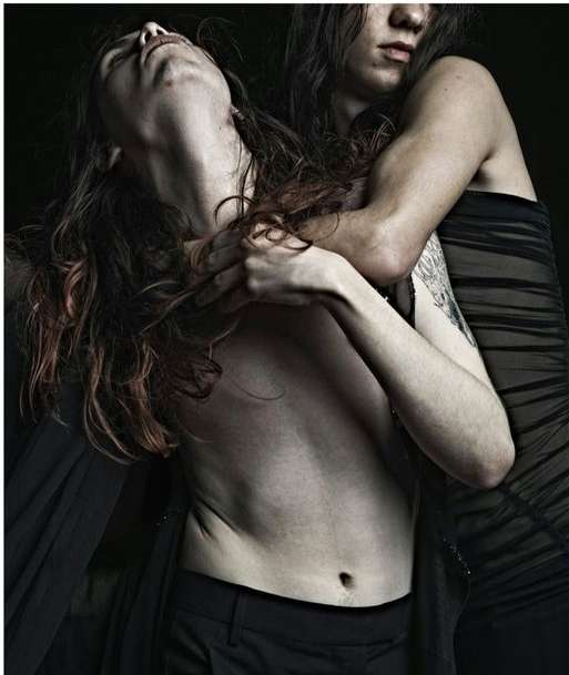 Two adults are closely embraced in a dimly lit, intimate setting. One person is shirtless and the other is in a dark garment.