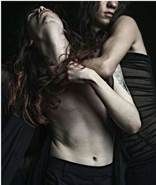 Two adults are closely embraced in a dimly lit, intimate setting. One person is shirtless and the other is in a dark garment.