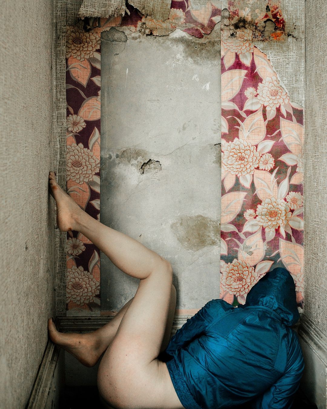 Woman in a hooded jacket reclines in a tight interior space with floral wallpaper and exposed concrete, legs raised against the wall.