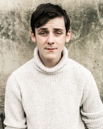 Close-up portrait of a young adult with dark hair wearing a white turtleneck sweater, against a textured grey grunge background.