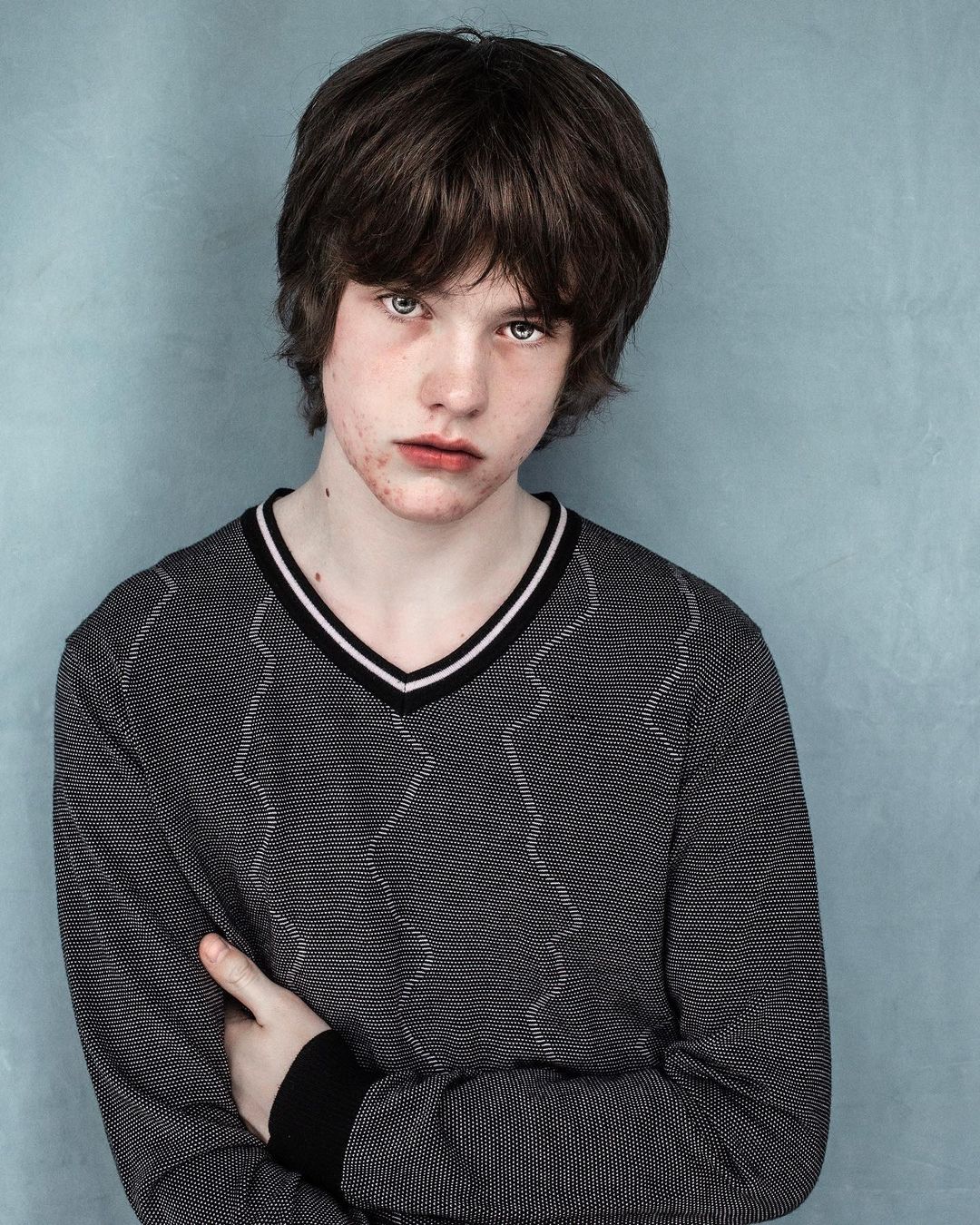 Portrait of a young adult with dark hair wearing a patterned V-neck sweater against a textured light blue wall backdrop indoors.
