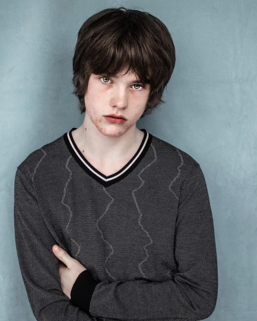 Portrait of a young adult with dark hair wearing a patterned V-neck sweater against a textured light blue wall backdrop indoors.
