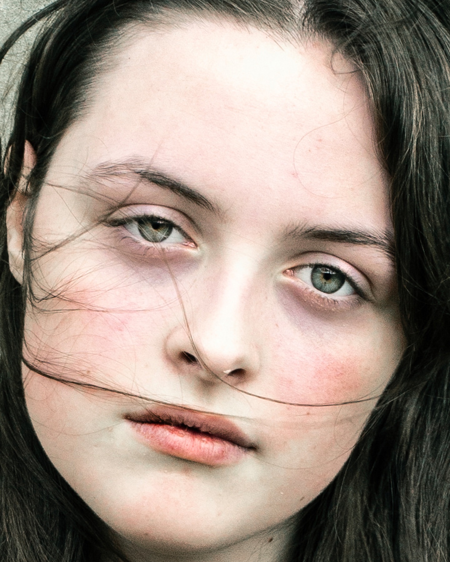 Close-up portrait of a young woman with fair skin, dark hair, and light-colored eyes. Her face is framed by loose strands of hair.