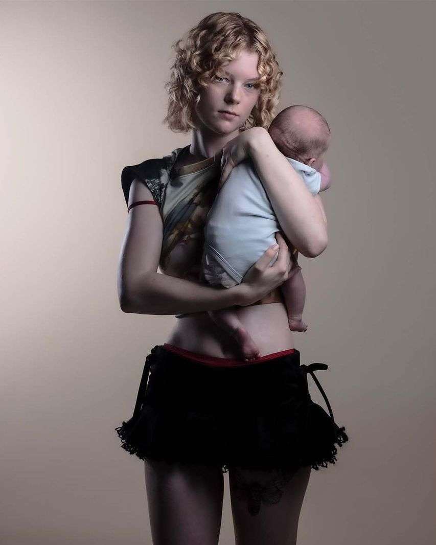 An adult female holds a newborn baby. The background is a neutral color. The female wears black shorts and a patterned top.