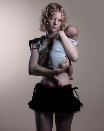 An adult female holds a newborn baby. The background is a neutral color. The female wears black shorts and a patterned top.
