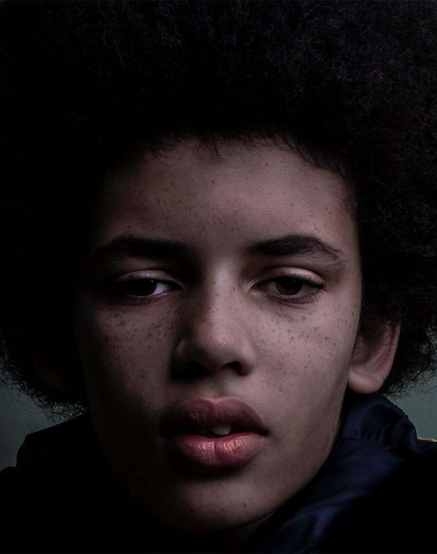 Portrait of a young person with dark, curly hair and freckles, looking directly at the camera. The lighting is dramatic and moody.