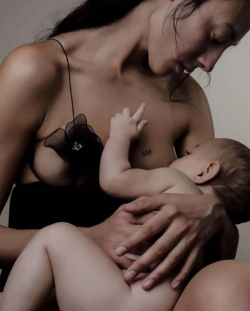 A mother nurses her infant in a close-up shot. The mother, wearing a black dress, looks down at her baby, the image is softly lit.